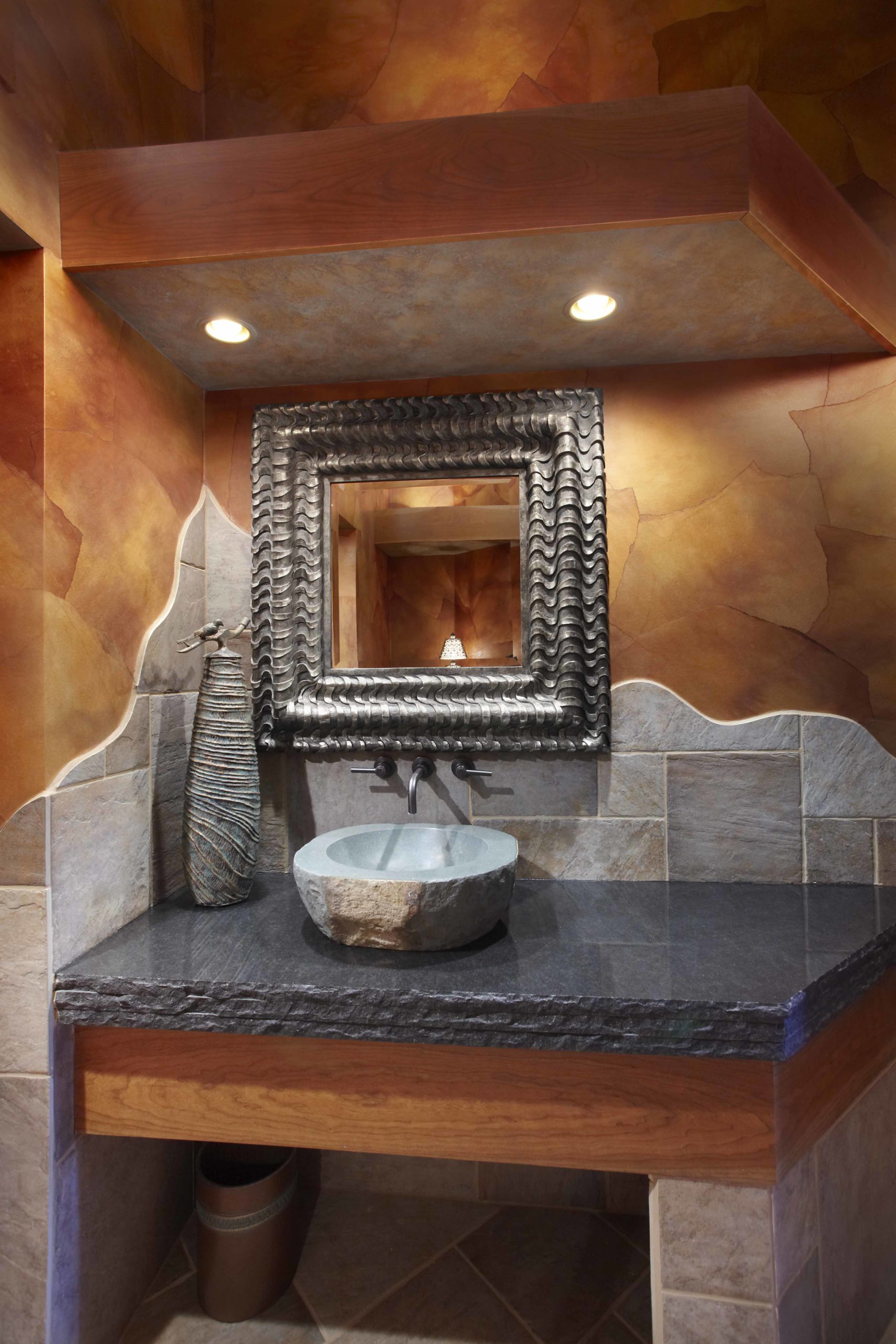Bathroom with brown and grey stone on the walls and a square mirror hanging over the vanity